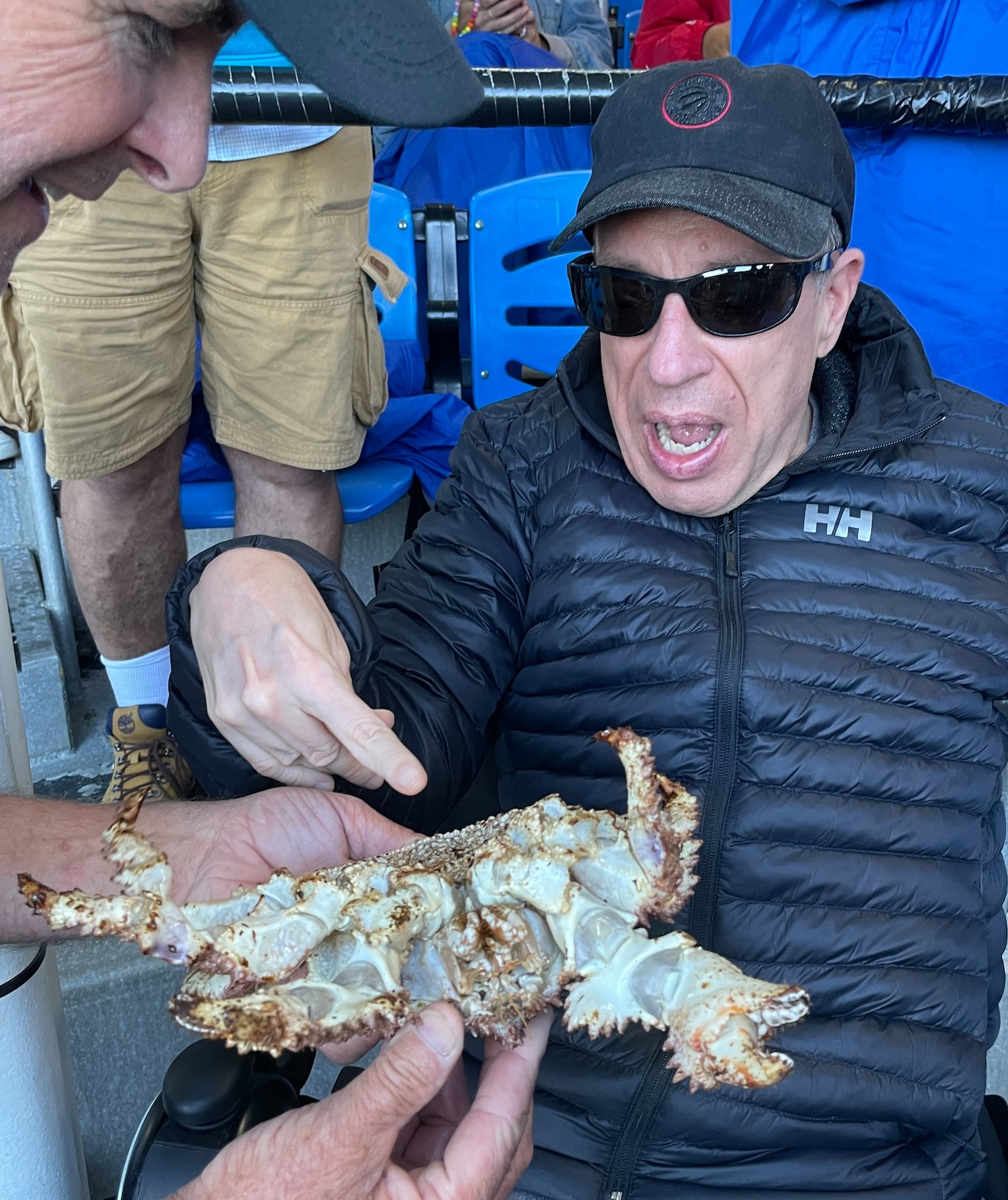 David, in a jacket, baseball cap, and sunglasses, looking shocked and pointing at a giant crab that someone off camera is holding out to him.