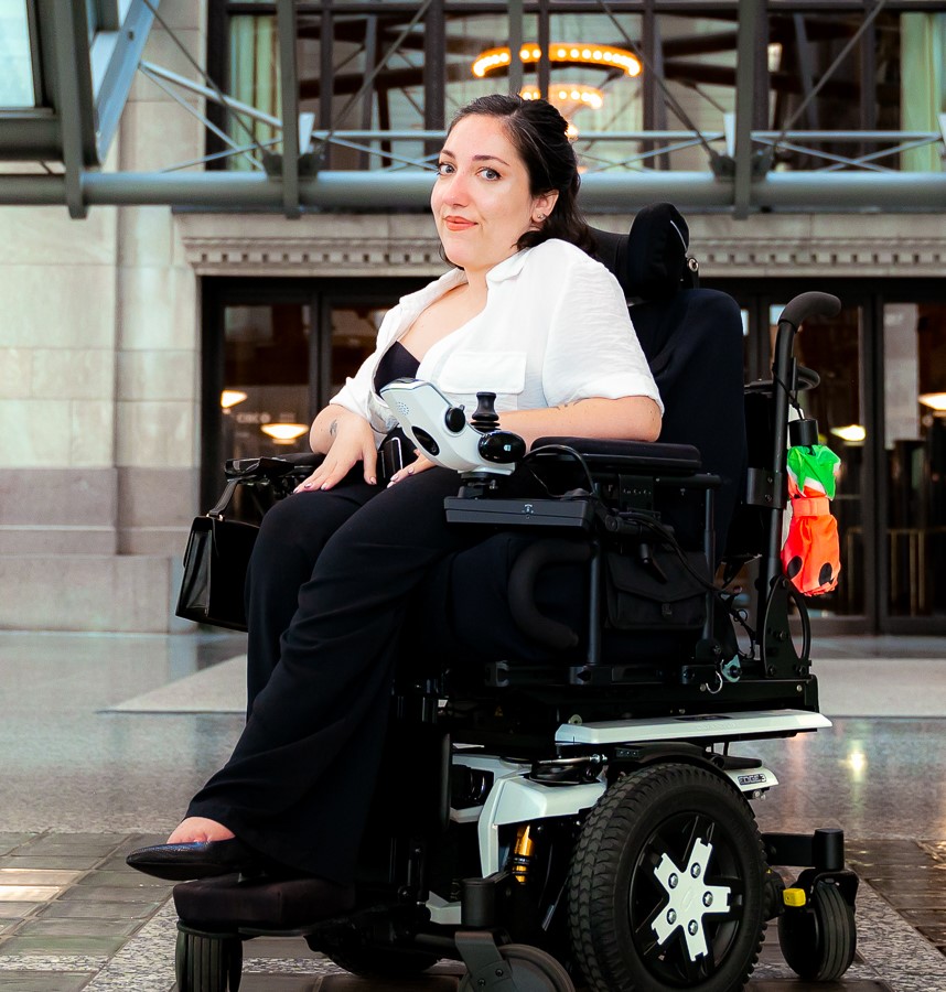 Jessica, seated in a mobility device, poses near an entryway.