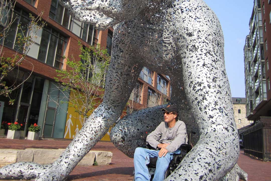 Man sitting in powered wheelchair under sculpture.