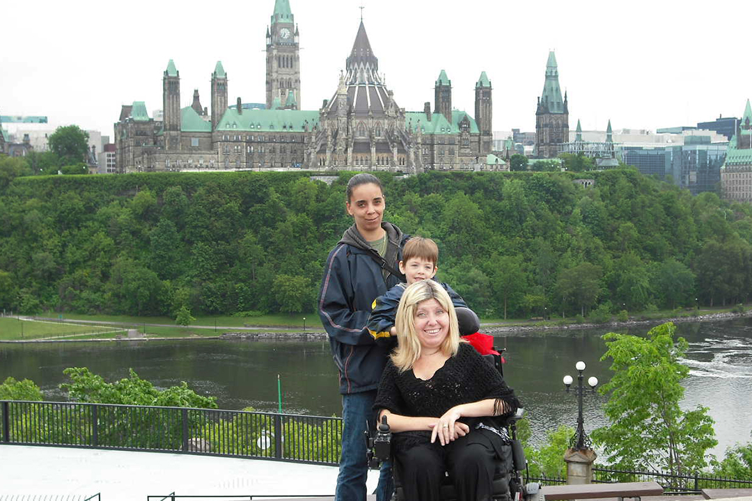Mother in wheelchair sitting in front of son and attendant, Parliament in the background.