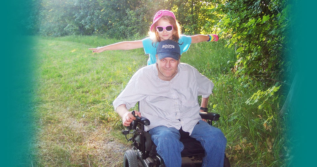 A man sitting in a powered wheelchair with his daughter riding behind him smiling in a forest clearing.