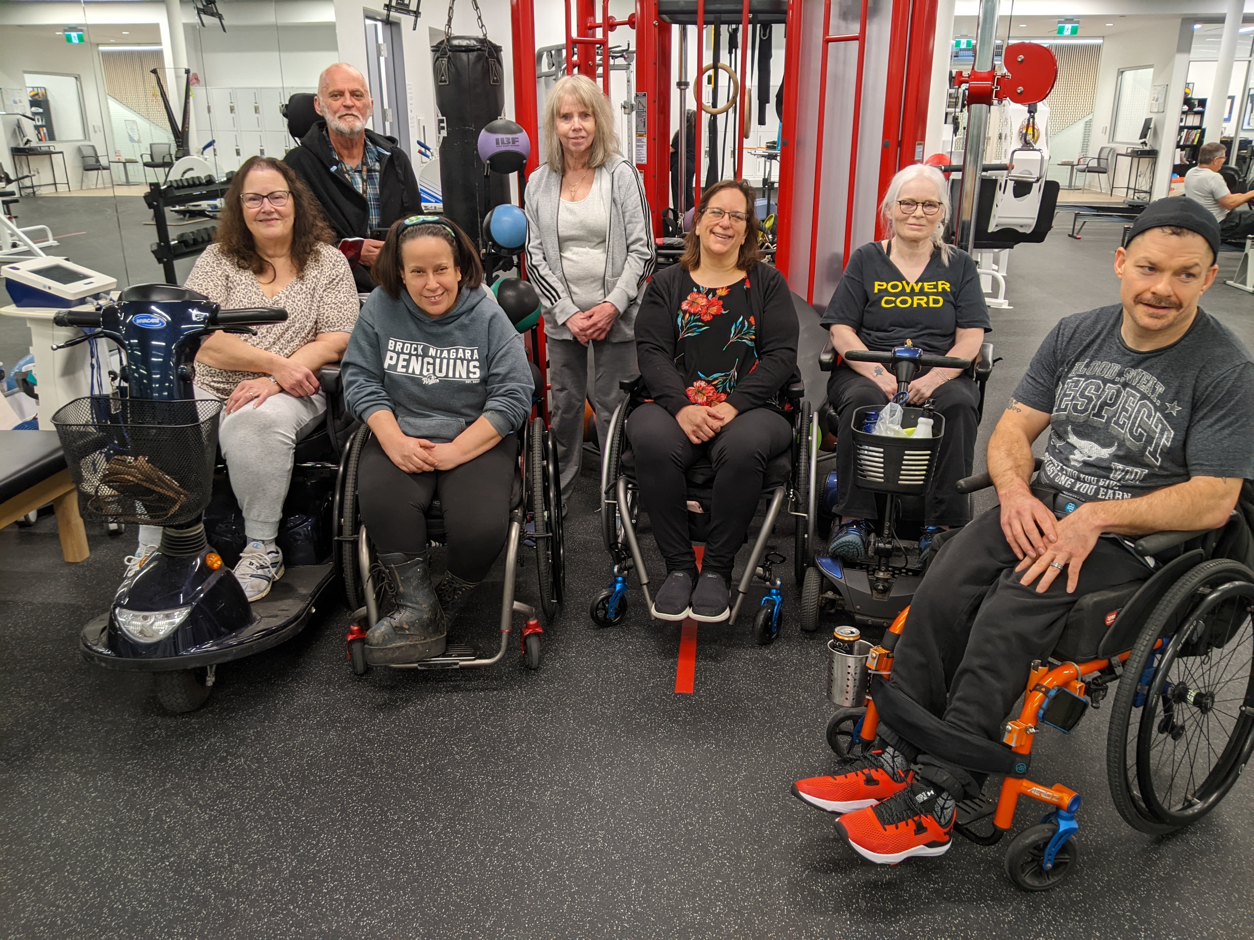 Seven individuals pose for the camera in two rows in front of gym equipment. Most are seated in mobility devices, and one is standing.