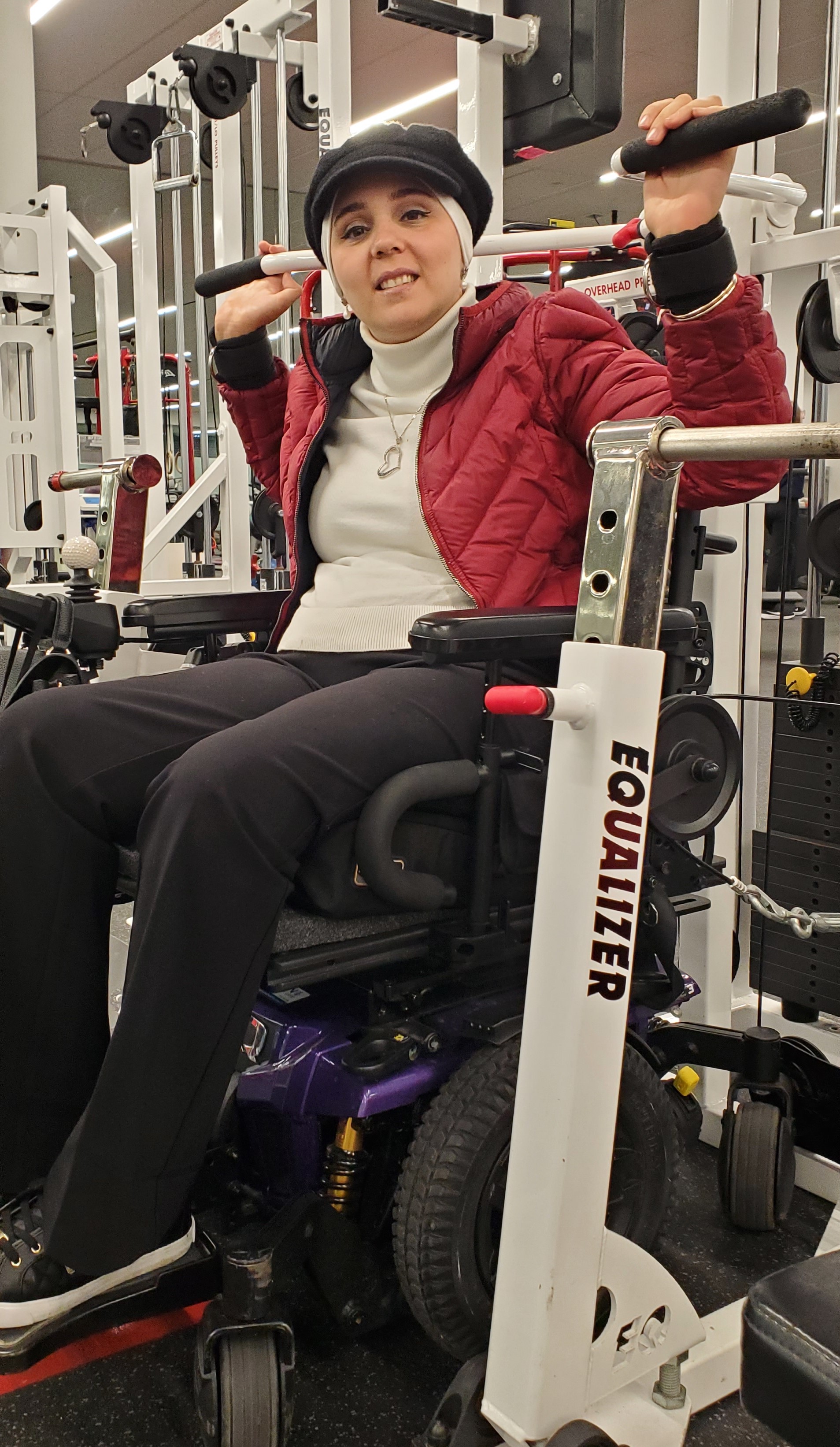 Amani, wearing a red jacket and black hat, is seated on the shoulder press machine. Her arms are up at head level on either side of her, gripping the handles of the machine.