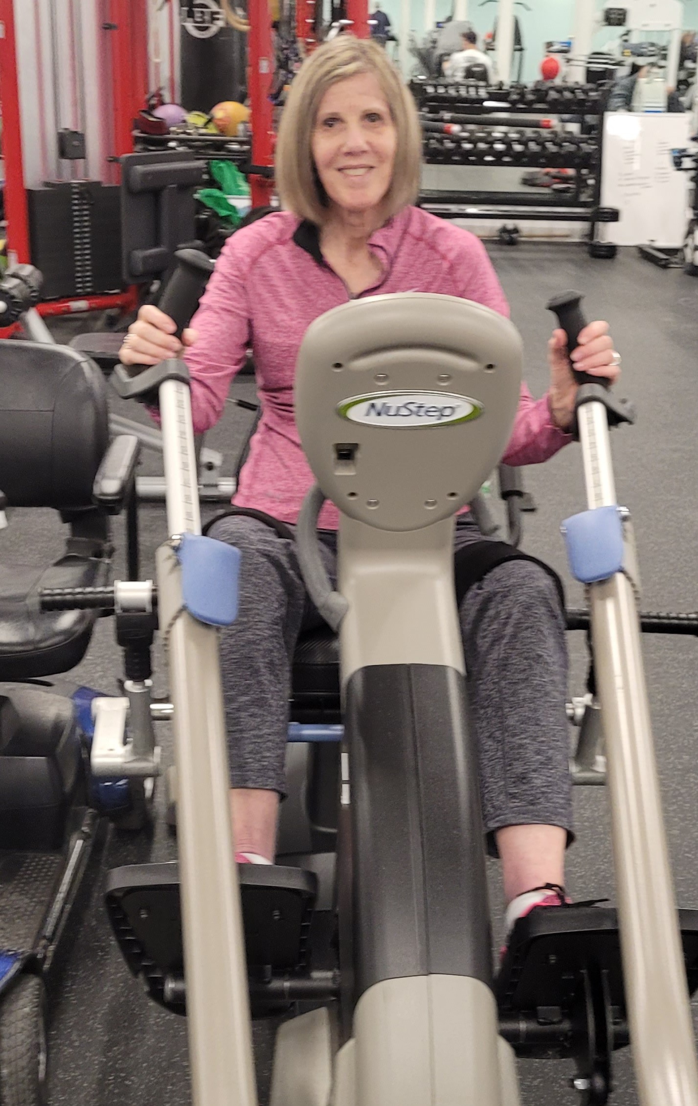 Sharon, wearing a pink top and grey pants, smiles while using a stationary bike.