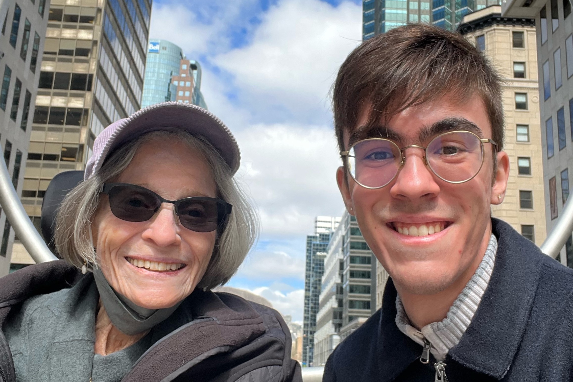 Dorothy and grandson Daniel from the shoulders up, smiling at the camera. The background is city buildings against a cloudy blue sky.