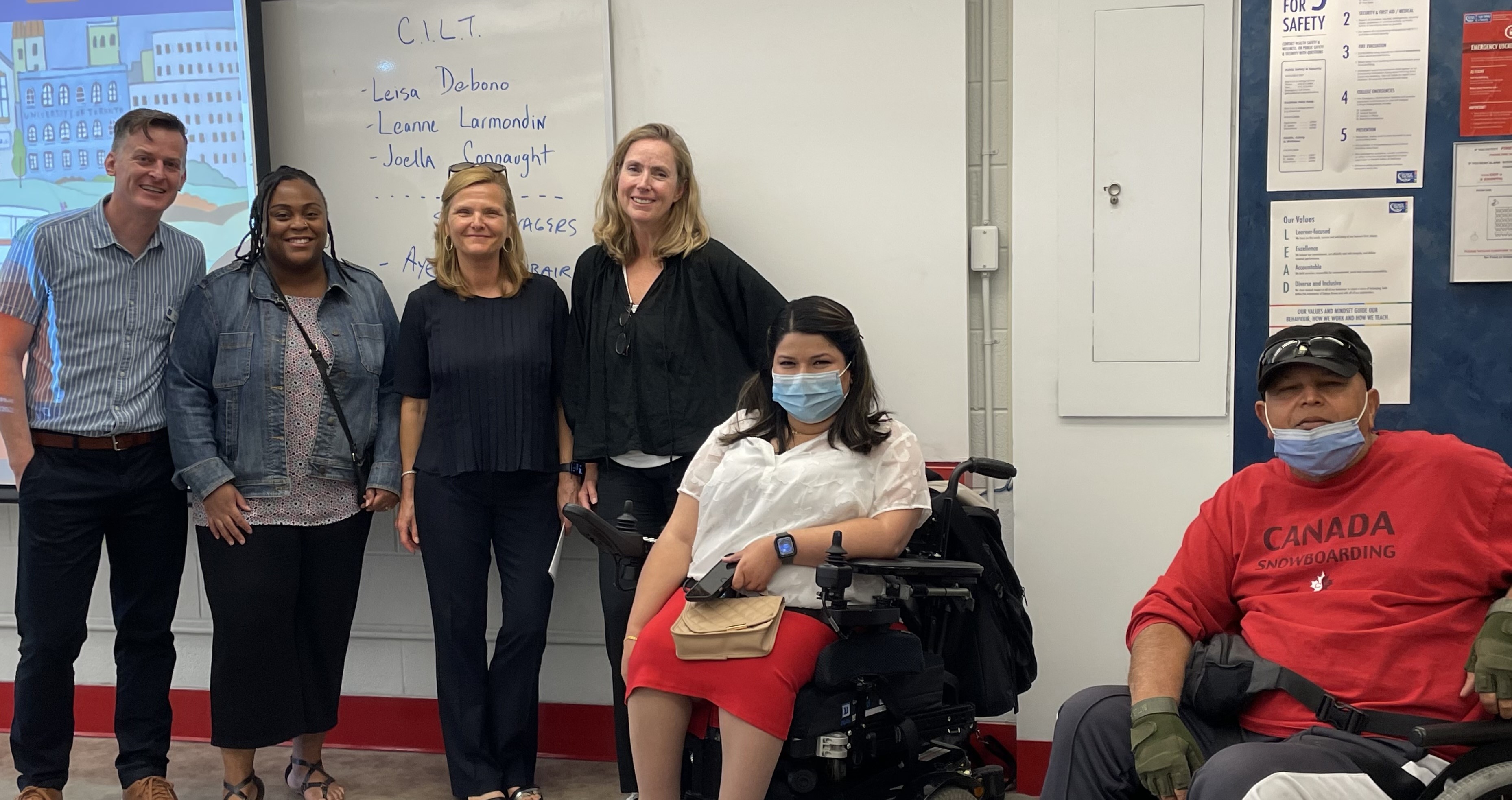 Presenters and professor stand or sit in front of a whiteboard at the front of a classroom. The board has their names on it.