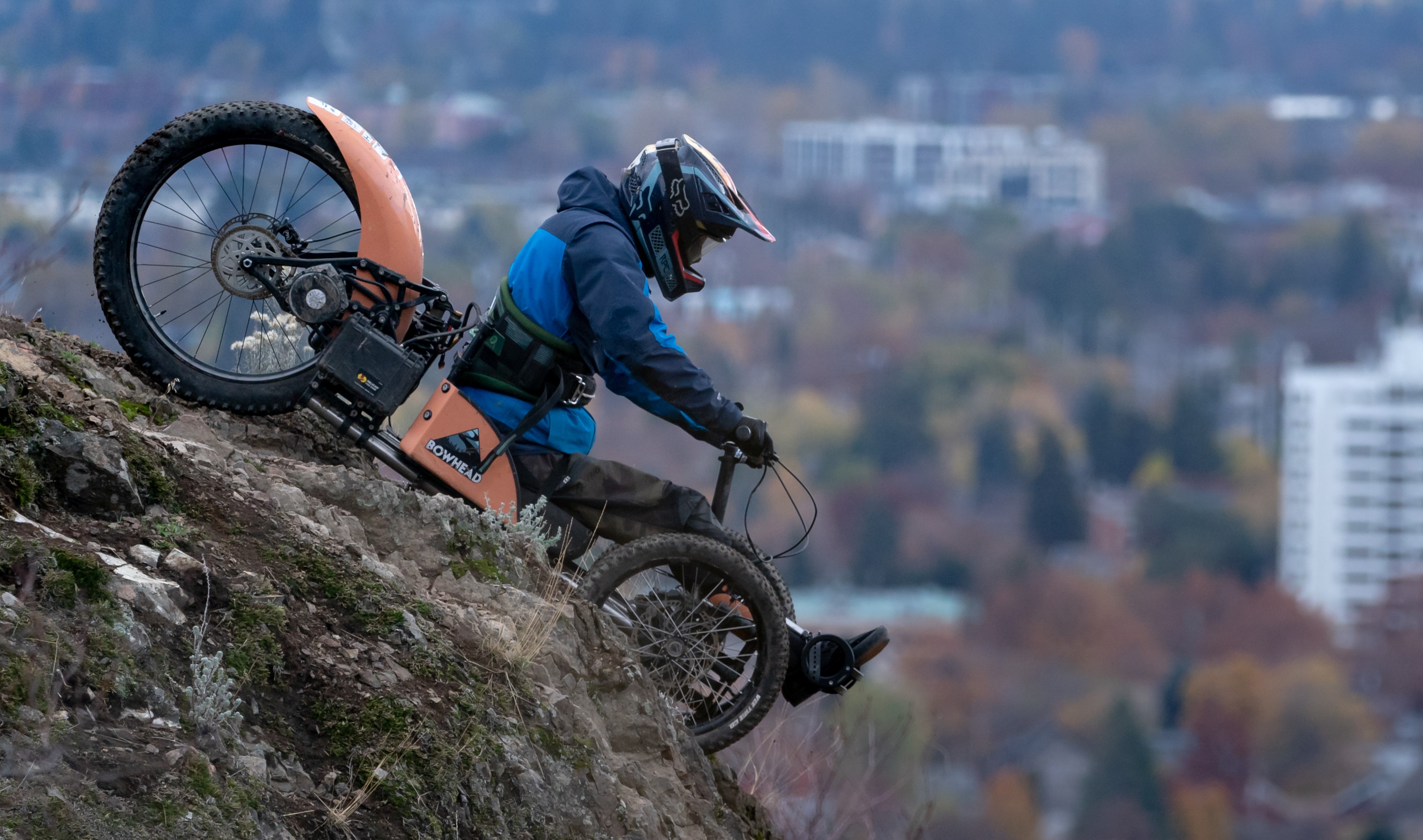 An adaptive bike leans at a dramatic 45-degree angle over the edge of a hill. The driver's face is obscured by a helmet.