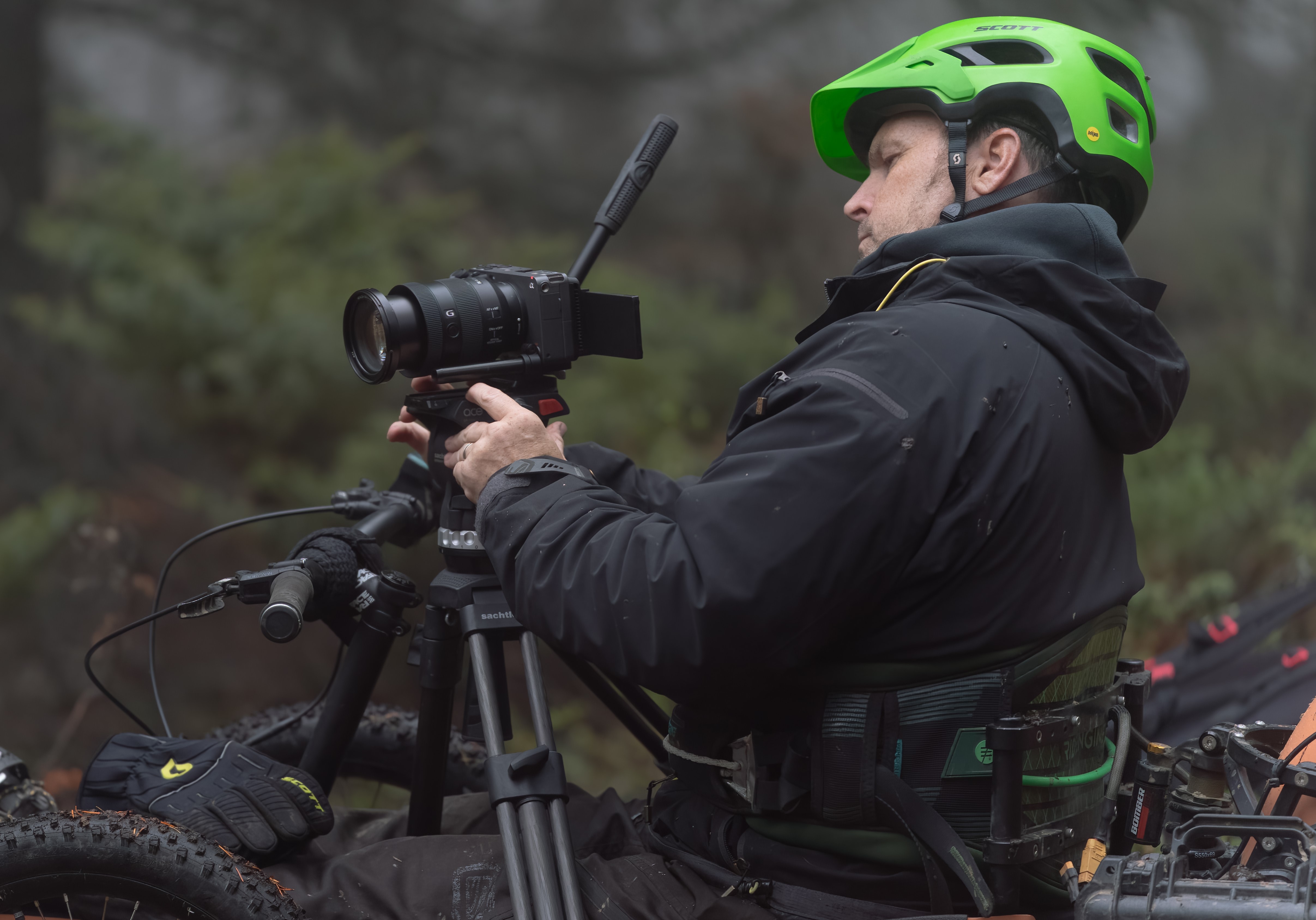 Jake, wearing a lime green helmet, films something off-screen with his camera on a tripod.