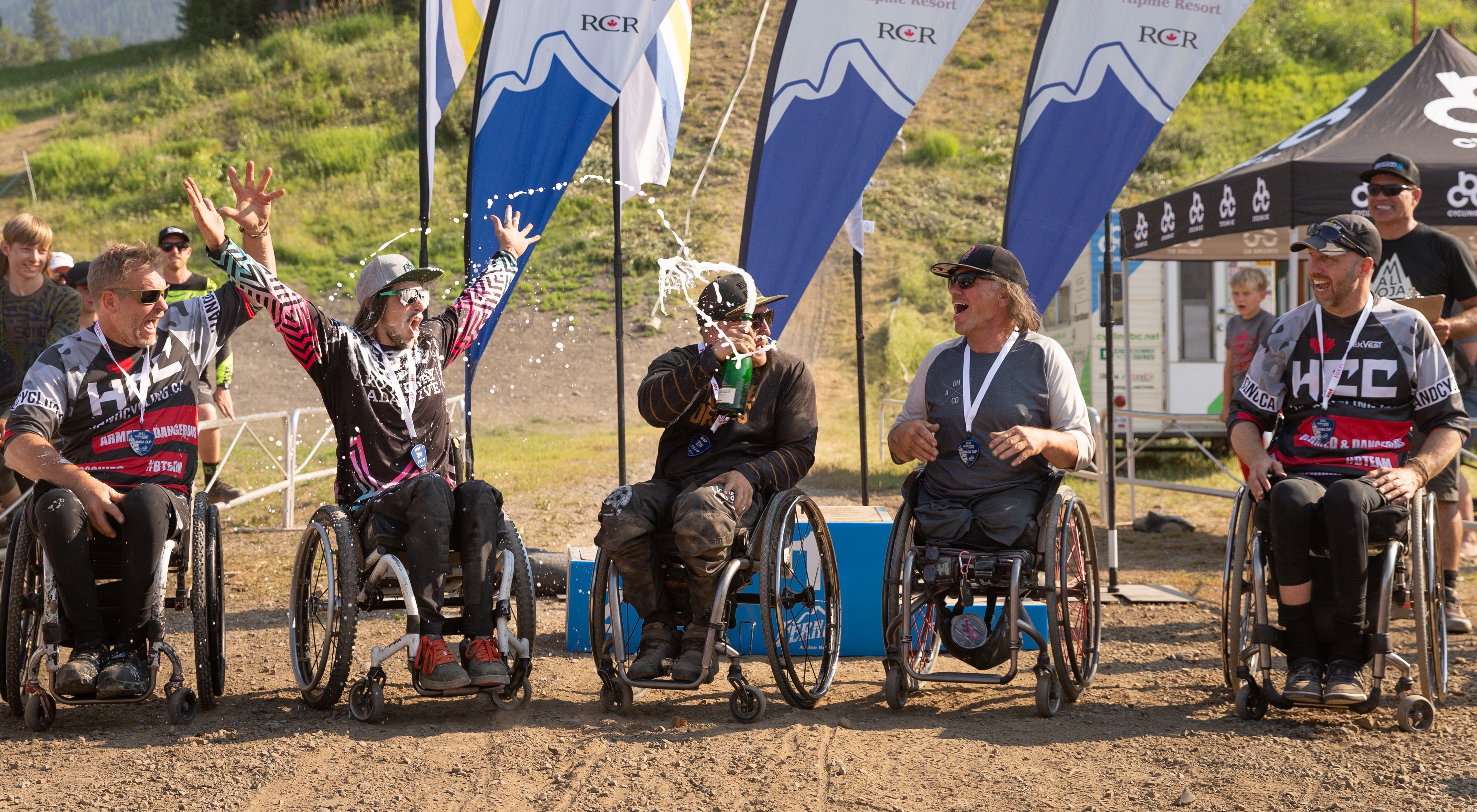 Five individuals are seated in wheelchairs in front of a blue podium and three flags that say 