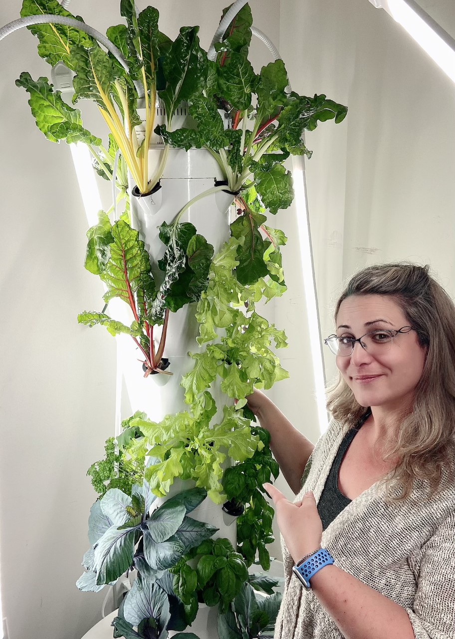 Jayme smiles and points at her tower garden, a vertical white structure with leafy green plans growing out of it at various points. Long lights extend out from tubes attached to the top of the structure.