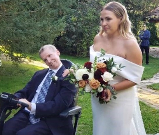 John, using a mobility device, beside his daughter, Amanda, who is standing in a wedding dress and holding a bouquet of flowers. The setting is outdoors in a green space.