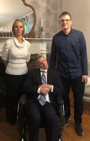 Two attendants stand on either side of John, who is seated and wearing a suit and tie. They are all posing for the camera.