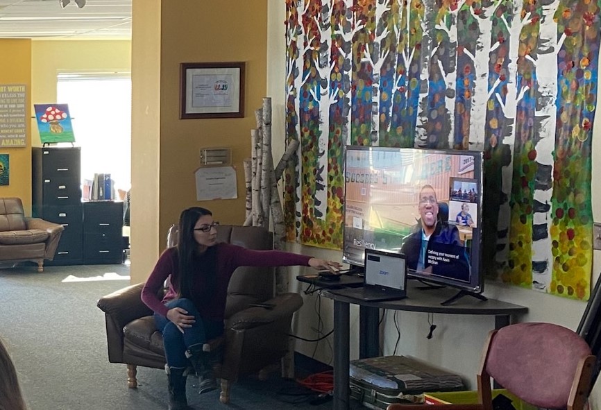 Georgina sits on a comfortable chair next to a TV and computer. She is reaching over to adjust something with the technology. Kevin is shown on the TV screen for his live presentation.