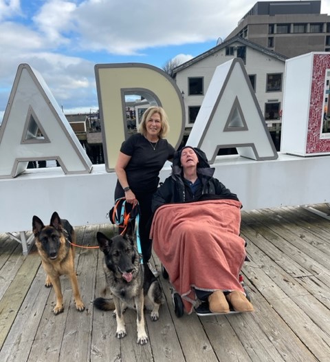 Mark Hardes, sitting in a mobility device under a blanket, poses with a standing woman holding the leashes of two large dogs. They are posing outdoors in front of a large 