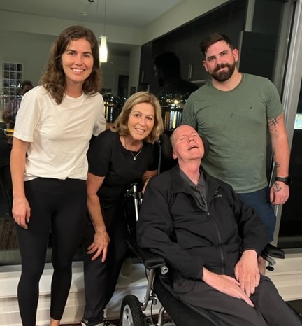 Mark, seated in a mobility device, poses with three other people standing behind and beside him. All are smiling.