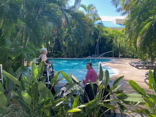 Mary Margaret and Stella sit at the side of a pool with greenery in the background. Stella is looking over her shoulder at the camera. There are leaves in the foreground.