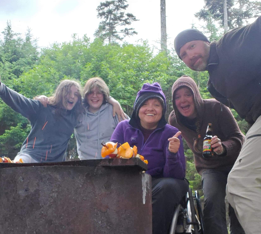 Five individuals, including Melissa Webster, are gathered outside and posing for the camera together while preparing skewars on a grill.