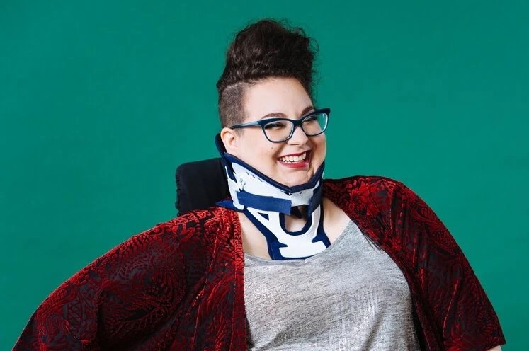 Shoulders-up photo of a smiling Ophira, who has their hair up and is wearing glasses and a neck brace.