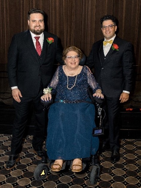 Penny Goldberg in a blue formal outfit. Two men in suits are standing behind her. All are smiling for the camera.