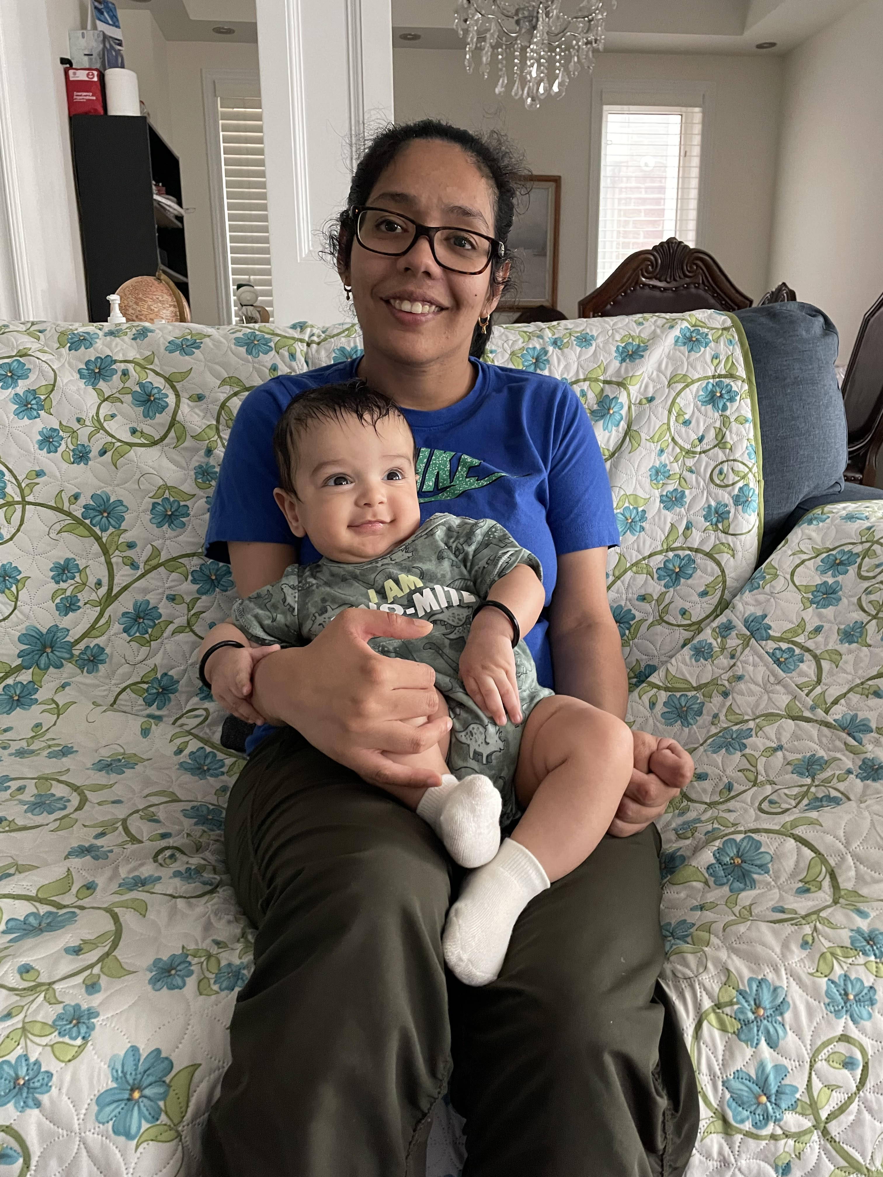 Priyanka holds baby Brij on her lap, while sitting on a floral couch.