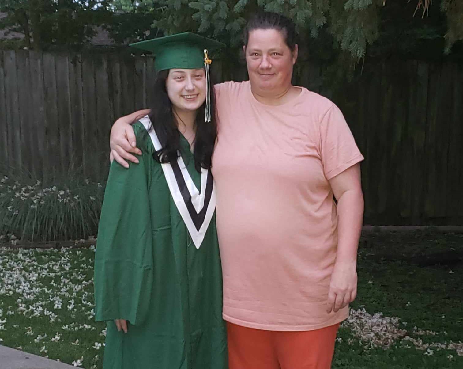Alexis and Amanda stand in a garden, with Amanda's arm wrapped around Alexis's shoulders. Alexis wears a green graduation gown and cap.