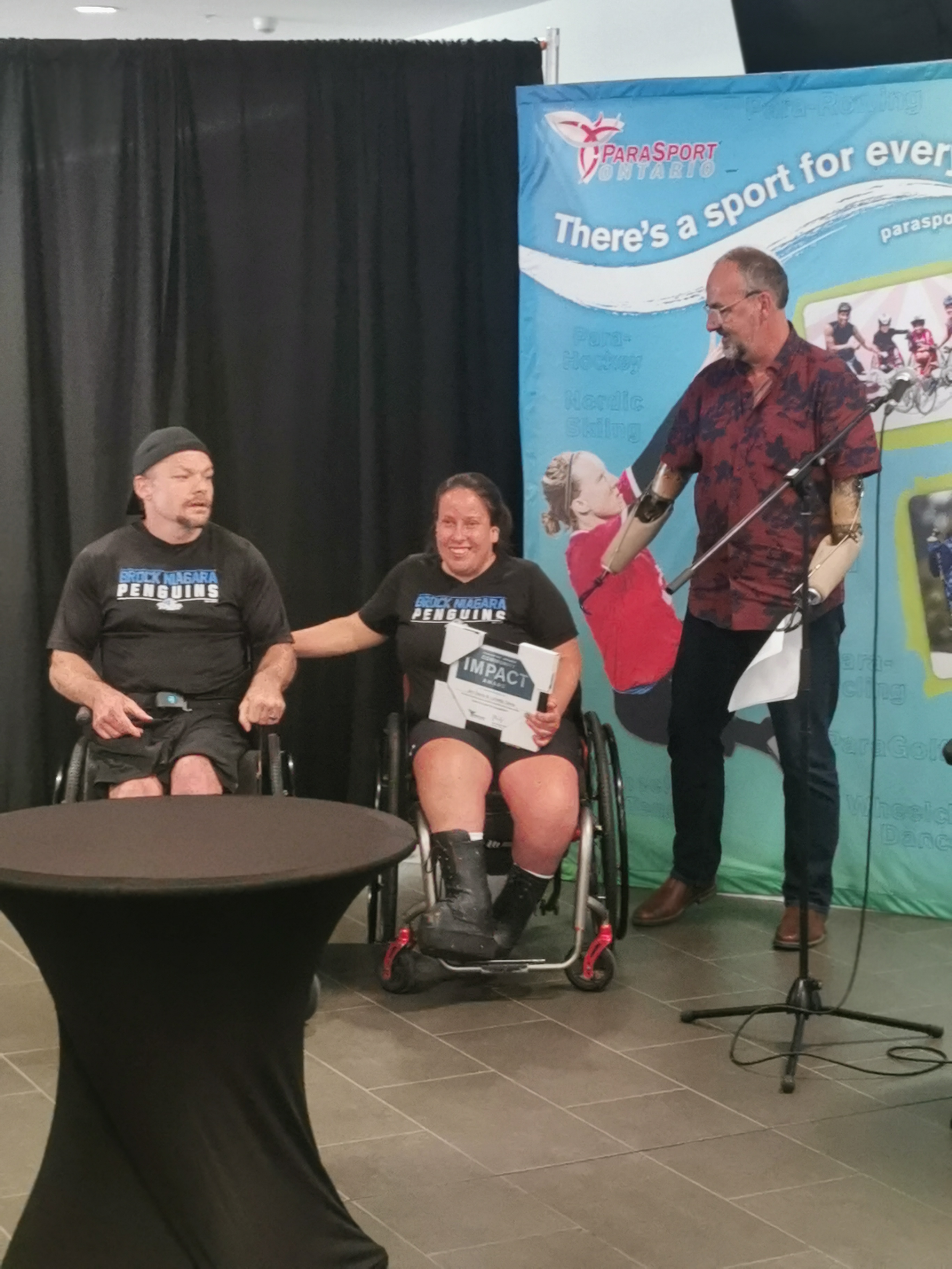 James and Loretta received an award from a presenter in front of a sports banner.