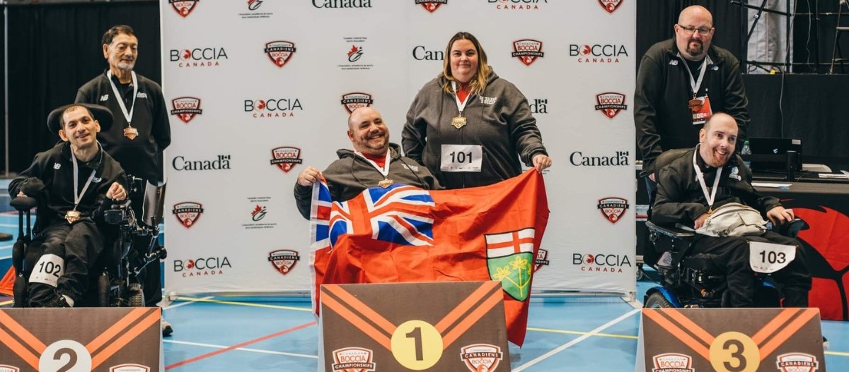 Three pairs at the podium with medals around their necks. The pair in the middle holds a large Ontario flag.